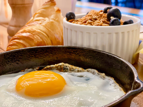 Eating breakfast food inside a caf&eacute; at the table, fried egg, yellow yolk served in a frying pan, cheese slices, croissant and fresh berries in a bowl. Depth of field blurred background. 