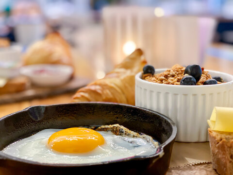Eating breakfast food inside a caf&eacute; at the table, fried egg, yellow yolk served in a frying pan, cheese slices, croissant and fresh berries in a bowl. Depth of field blurred background. 