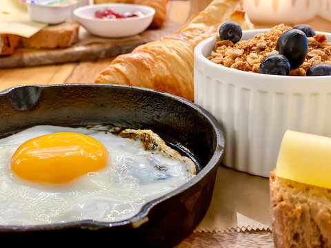 Eating breakfast food inside a caf&eacute; at the table, fried egg, yellow yolk served in a frying pan, cheese slices, croissant and fresh berries in a bowl. Depth of field blurred background. 