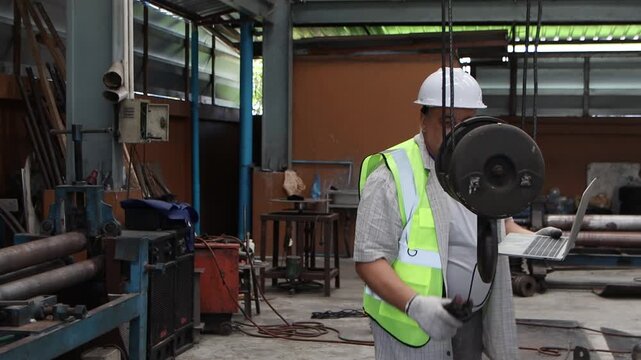 Industrial senior engineer in safety vest and white helmet holds laptop to inspect machinery at manufacturing metal factory. Senior foreman maintains lifting machine and large steel hook at factory.