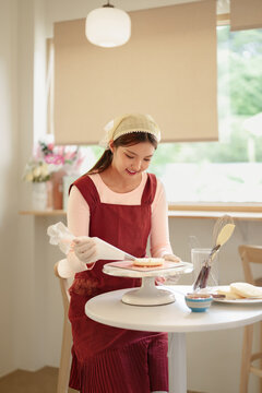 Young Asian woman decorating homemade cake on stand in bright modern kitchen with copy space