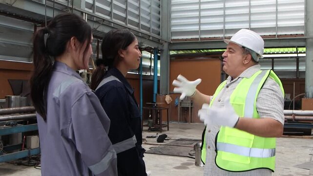 Happy elderly engineer supervisor with safety vest and helmet praising, encouraging two Asian female workers. Senior foreman offers compliment, pats on shoulder and friendly hug with staffs at factory