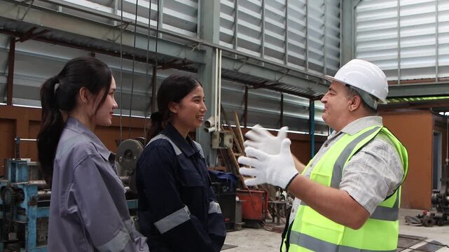 Happy elderly engineer supervisor with safety vest and helmet praising, encouraging two Asian female workers. Senior foreman offers compliment, friendly pat on shoulder from industrial factory staffs.
