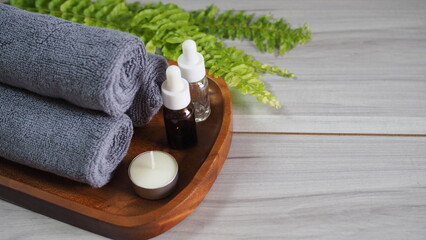 Minimalist spa still life with rolled dark gray charcoal towels, essential oil dropper bottles, and a tea light candle on an organic-shaped wooden tray. Decorated with green fern leaves on a light gra © kanjana