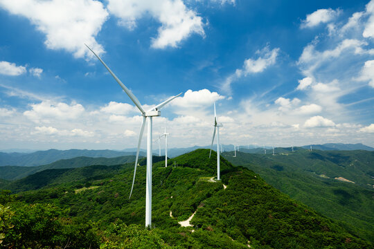 Aerial View of Wind Farm on Green Mountain Ridges Under Blue Sky and Clouds, Multiple Wind Turbines Generating Renewable Energy Across a Lush Forested Landscape