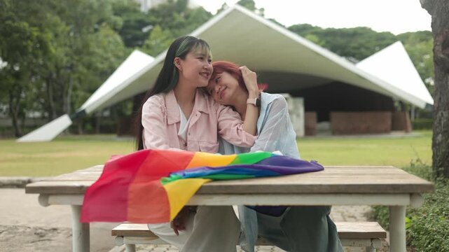 Two gentle Asian women sitting at a table in the park, one softly resting her head on her partner's shoulder under a vibrant rainbow LGBTQ pride flag, displaying intimacy and true romance.