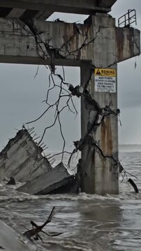 Pier ruins coastal warning sign visible, collapsed deck slabs and jagged rebar teeth rise from churning tide, concrete pillars pounded and undercut by waves, rust streaks and exposed steel, foam