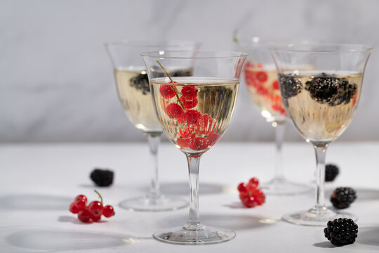 Crystal glasses filled with sparkling champagne sit on a light-colored table. The champagne glasses contain various berries. The light casts shadows through the glasses.