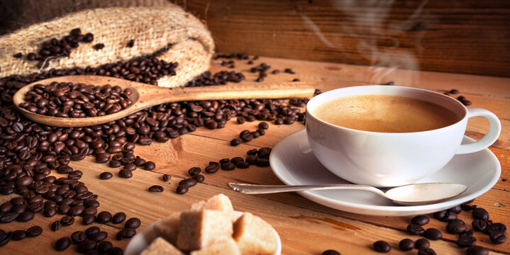 Cup of hot coffee and freshly roasted coffee beans with jute sack on rustic wooden work surface. Close-up.