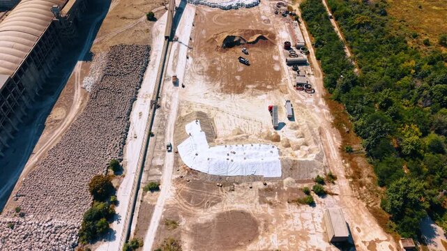 Raw material storage yard at cement factory. Top down aerial view of aggregate piles, covered stock, dump trucks, concrete parts and trees along the site.