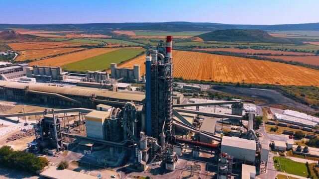 Cement production tower surrounded by harvested fields. Aerial perspective of a tall kiln structure, conveyors, silos and factory buildings within patchwork farmland.
