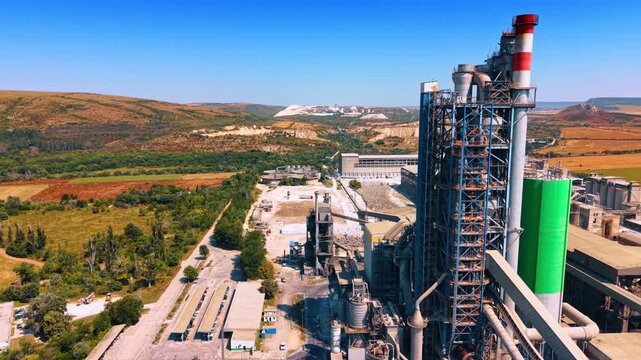 Cement plant preheater tower beside open quarry. Aerial view of steel framework, striped stack, green silo, service roads and excavated hills under blue sky.