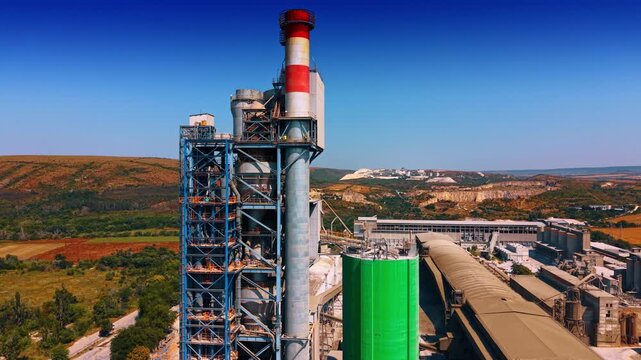 Cement preheater tower with striped industrial stack. Close aerial view of blue steel framing, red and white chimney, green silo, ductwork and factory roofs.