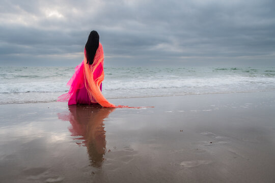 art portrait of anonymous woman in long pink dress in ocean shore wrapped in thin orange fabric