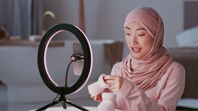 Focused Young Asian Woman Demonstrating Hand Knitted Socks While Sitting In A Cozy Home Environment With A Ring Light, Emphasizing Craftsmanship And Practicality. Healthcare Concept.