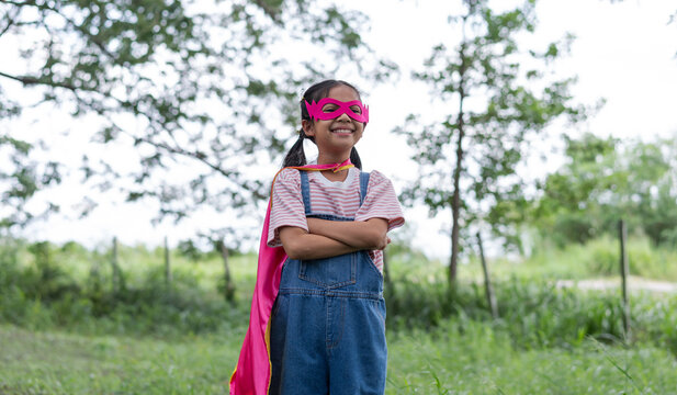 A confident young Asian girl smiles with arms crossed in a park. She wears a homemade pink superhero mask, cape, and denim overalls, showing girl power and imagination outdoors.