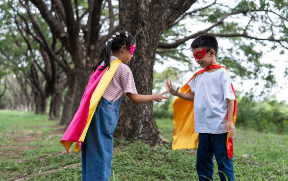Two Asian children in homemade superhero costumes play a rock paper scissors game outdoors. The girl and boy enjoy a fun competition, showing strategy and sibling teamwork in a park.