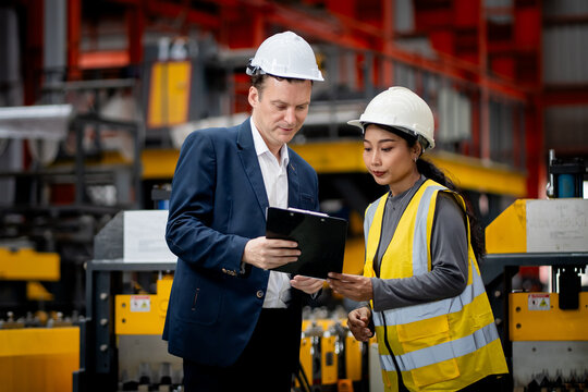Two professional engineer,worker,technician use clipboard discuss work, walk in steel metal manufacture factory plant industry. Black African American man and woman wear hard hat check quality machine