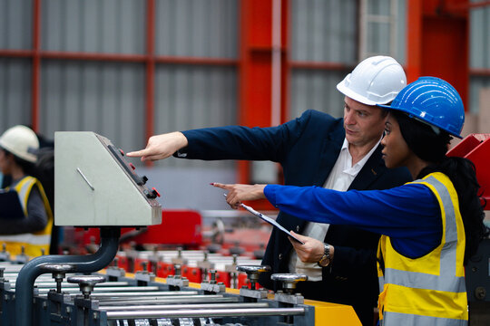 Two professional engineer,worker,technician use clipboard discuss work, walk in steel metal manufacture factory plant industry. Black African American man and woman wear hard hat check quality machine