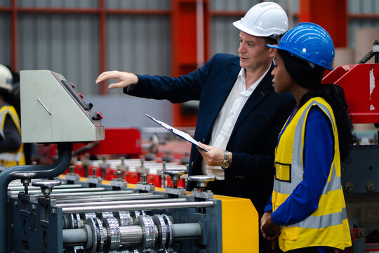 Two professional engineer,worker,technician use clipboard discuss work, walk in steel metal manufacture factory plant industry. Black African American man and woman wear hard hat check quality machine