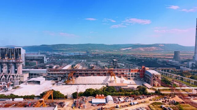 Industrial plant area with white mineral piles and conveyor machinery. Aerial view of chemical factory site, raw material heaps, processing structures, hills under clear sky.