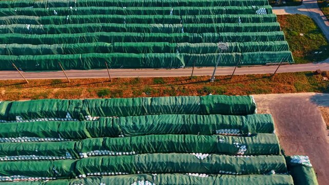 Industrial logistics depot, long rows of covered supply cargo piles. An aerial view captures green protective sheets over large piles of bags next to an asphalt road.