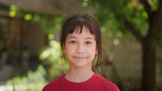 Portrait of adorable Asian kid looking directly at the camera with a gentle smiling face in a red shirt standing outdoors with beautiful green bokeh background. Childhood happiness lifestyle concept.