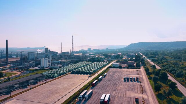 Industrial plant with cargo trucks and storage area under clear sky. An aerial view shows a large factory complex with parked semi vehicles and rows of green covered materials.