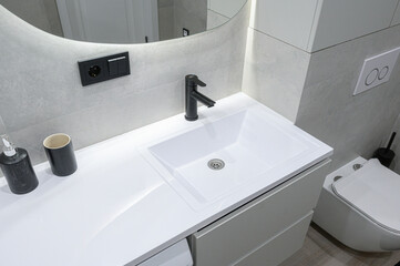 Fototapeta premium modern bathroom features a white integrated rectangular sink with a black faucet, a curved mirror, minimalist accessories, a wall‑mounted toilet, and light‑gray tiled walls