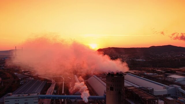 Factory buildings covered in heavy smoke at sunset. Dense orange and white vapor rises from a round tower structure over an industrial manufacturing site.