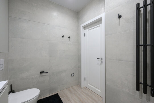 compact modern bathroom features a white sink with a black faucet, a wall‑mounted toilet, light‑wood flooring, gray tiles, black bath mats, black towel radiator for a sleek, minimalist look