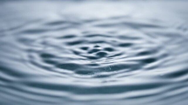 High speed macro photography of a water droplet impact creating a symmetrical crown splash with distinct spikes and a mirror reflection on a calm blue liquid surface.