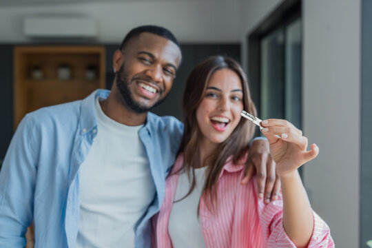 Cheerful young multiracial couple smiling widely while presenting a new house key, celebrating their first property ownership and successful real estate investment