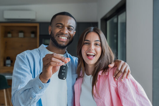 Smiling interracial couple standing in a bright modern apartment, proudly holding up a car key and celebrating a new vehicle or milestone together, conveying joy, excitement and togetherness.