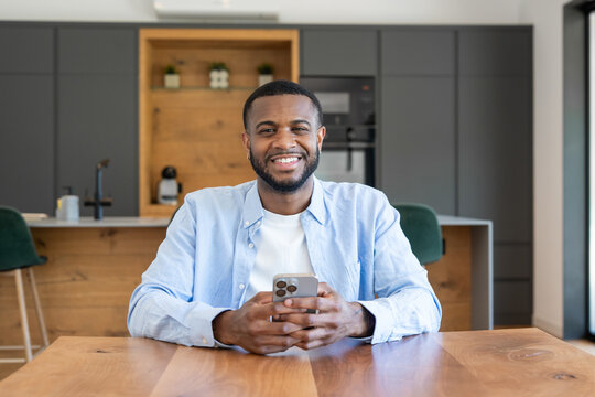 Smiling man sits at a wooden kitchen table holding a smartphone, relaxed and engaged with his device. Modern home interior suggests remote work, casual lifestyle, connectivity and positive mood.