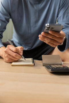 Hand smartphone notebook pen writing calculator desk finance business planning, man checks smartphone while writing notes, calculator and wallet on wooden table in office workspace Vertical