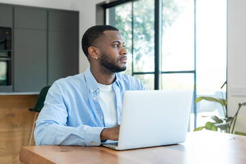Fototapeta premium Young black man working remotely on a laptop at a wooden table in a brightly lit modern home, focusing on tasks and contemplating new ideas for his business