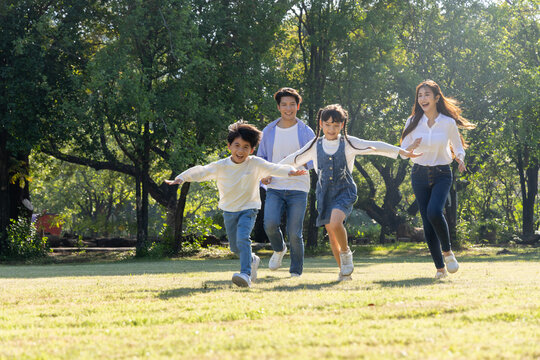 Happy Asian family with parents and young children running together on a green lawn in a sunny park. Enjoying outdoor activities, a healthy lifestyle, and quality time during summer leisure