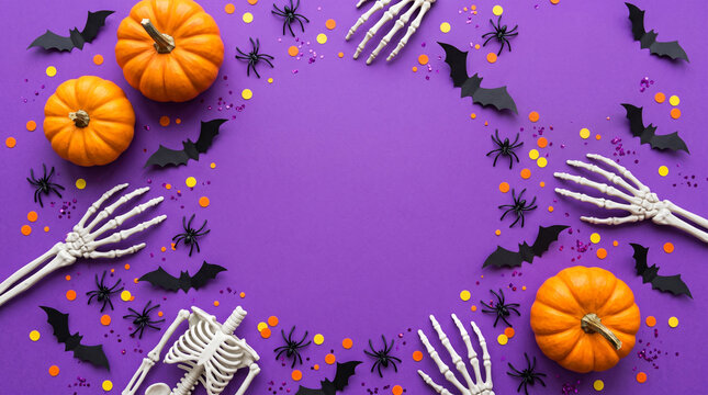 Halloween background with pumpkins, skeleton hands, bats, and spiders on a purple surface with confetti