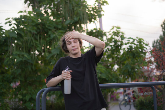 A young man stands near a railing with a bottle while drying his forehead in a park as the sun sets