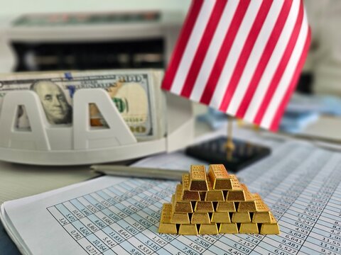Gold bars stacked on spreadsheets with a dollar bill and a small American flag
