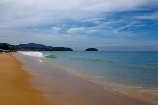 Tropical beach view with incoming waves on the sand and a lush green island coastline under cloudy sky