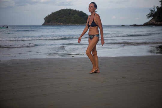 Young female traveler in swimsuit and hat looking back on a beautiful turquoise tropical sea beach with blowing blue cape