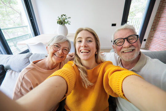 Young woman sits between her smiling grandparents and takes a cheerful selfie on a bright, cozy living room sofa, capturing family togetherness, warmth and multi-generational bonding.