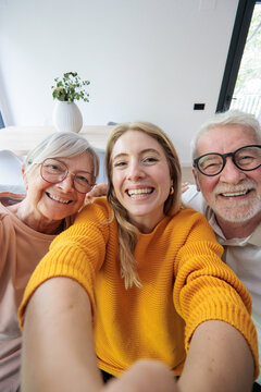 Young woman embraces her grandparents while taking a joyful selfie in a bright home, capturing genuine smiles, family connection and multigenerational warmth in a cozy living room moment.