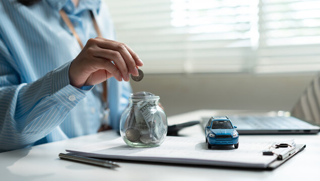 Woman hand putting coin into glass jar with paper money and blue toy car on contract document. Concept of saving money for buying new car, auto loan, investment, and insurance.