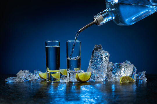 Shot glasses of vodka with lime slices and ice on a dark blue background.