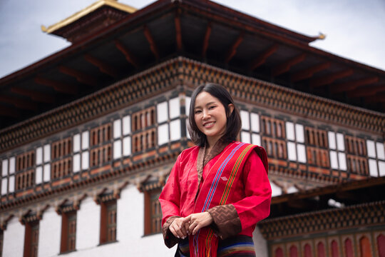 A happy young Asian woman in traditional red Bhutanese Kira dress, smiling warmly outside a fortress in Bhutan