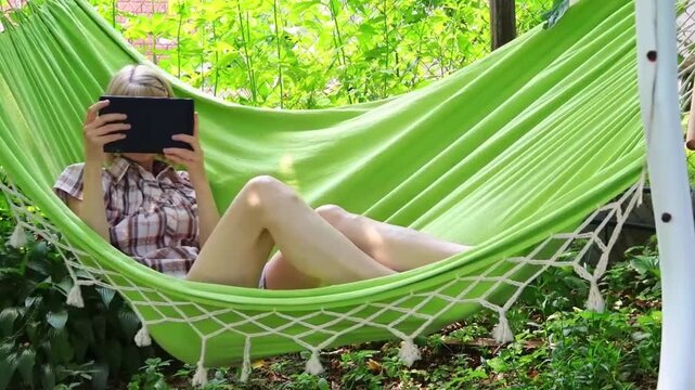 Young woman relaxing in bright green hammock while holding black tablet, surrounded by lush greenery, peaceful outdoor retreat, summer leisure time.