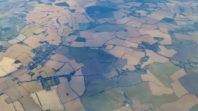 Aerial view of patchwork farmland landscape with varying shades of green and brown fields, winding roads, and small villages visible from above in a rural setting